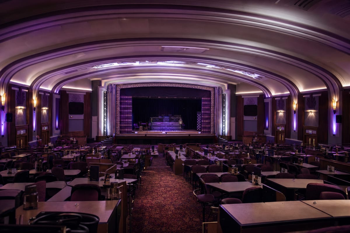 An empty bingo hall inside an old art deco cinema, with rows of tables leading to a stage under a curved art deco ceiling lit in violet (AI-generated image)