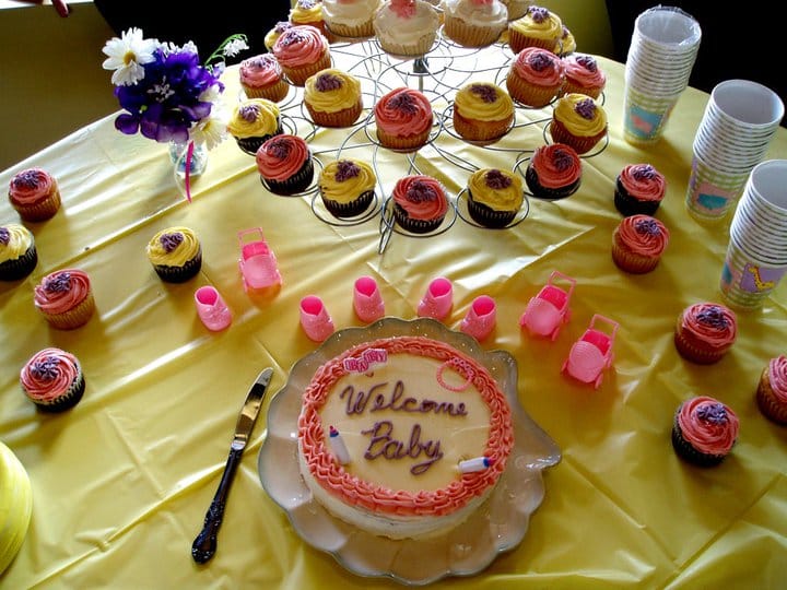 A table with a yellow cloth, a cake iced with Welcome Baby and a stand of pink and yellow cupcakes