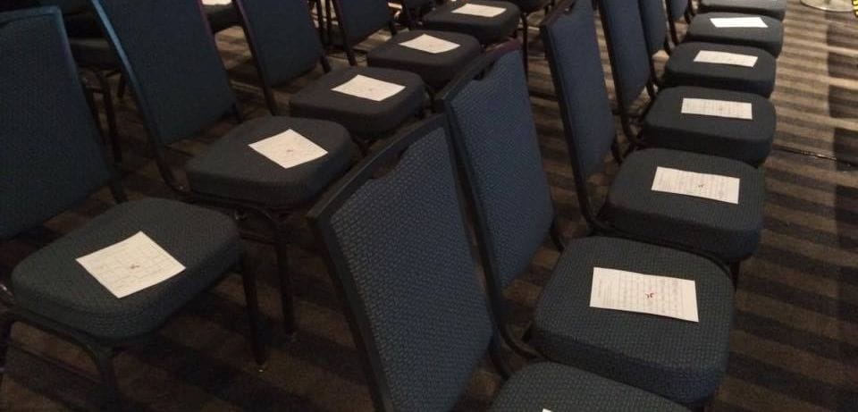 Rows of dark conference chairs, each with a printed bingo sheet on the seat